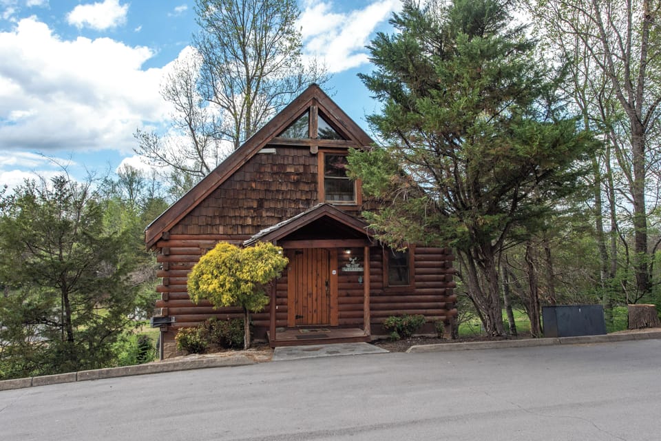 A charming log cabin retreat nestled among towering trees, featuring rustic wood siding and convenient parking for a perfect mountain getaway.