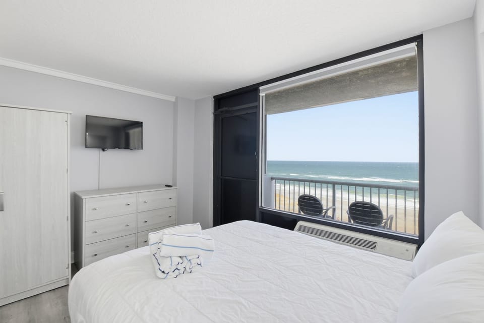 The cozy sleeping space invites relaxation with its tranquil color palette, plush bedding, and a panoramic view of the ocean, ideal for unwinding after a day at the beach.