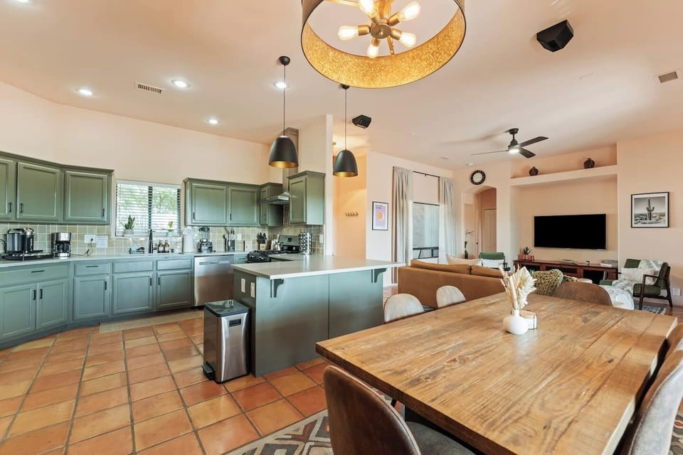 Spacious open-plan living area with modern kitchen, cozy dining space, and inviting lounge! Features stylish green cabinetry, a large wooden table, and a big TV. Perfect for family gatherings or entertaining, with warm lighting and terracotta floors!