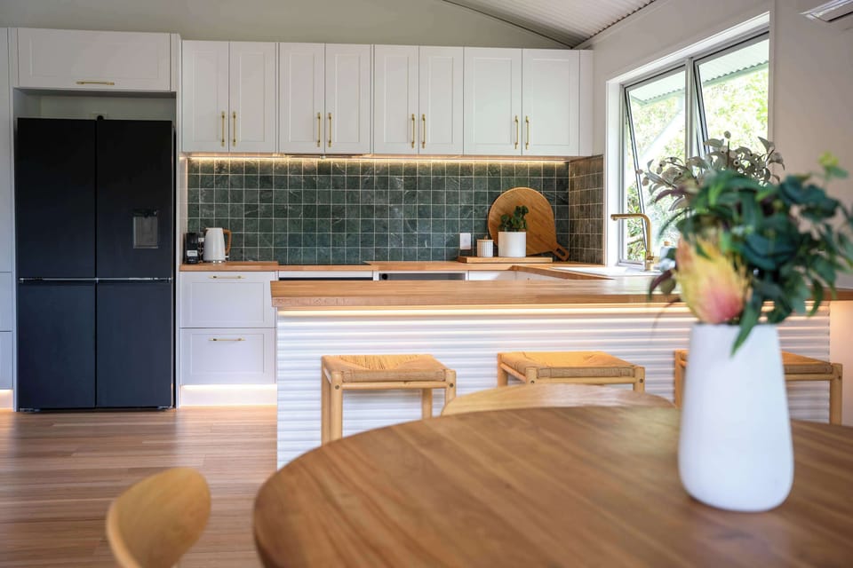 A bright and beautifully styled kitchen with timeless white cabinetry, warm timber finishes, and elegant gold accents ✨ The green tiled backsplash adds a fresh pop of color, creating a space that’s both functional and effortlessly inviting.