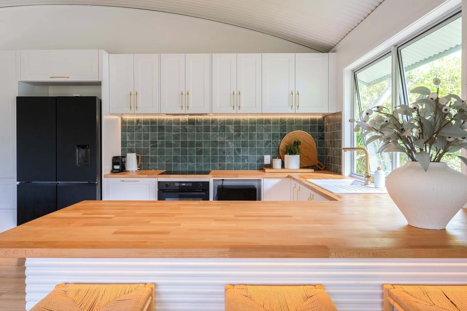 A bright and beautifully styled kitchen with timeless white cabinetry, warm timber finishes, and elegant gold accents ✨ The green tiled backsplash adds a fresh pop of color, creating a space that’s both functional and effortlessly inviting.