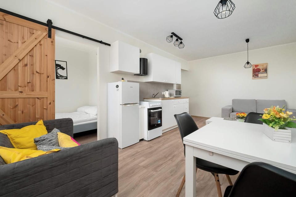 A bright open-plan living room and kitchen with white cabinets and wooden floors. A gray sofa with yellow accent pillows adds color to the modern space. The kitchen appliances are neatly integrated along the wall.