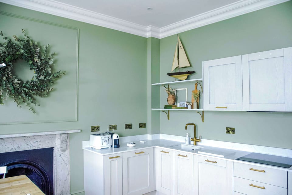 A bright, stylish kitchen with crisp white cabinetry, gold accents, and a sleek sink and faucet. Open shelving adds charming décor, while the space flows seamlessly into a warm dining area with a wooden table and white chairs.