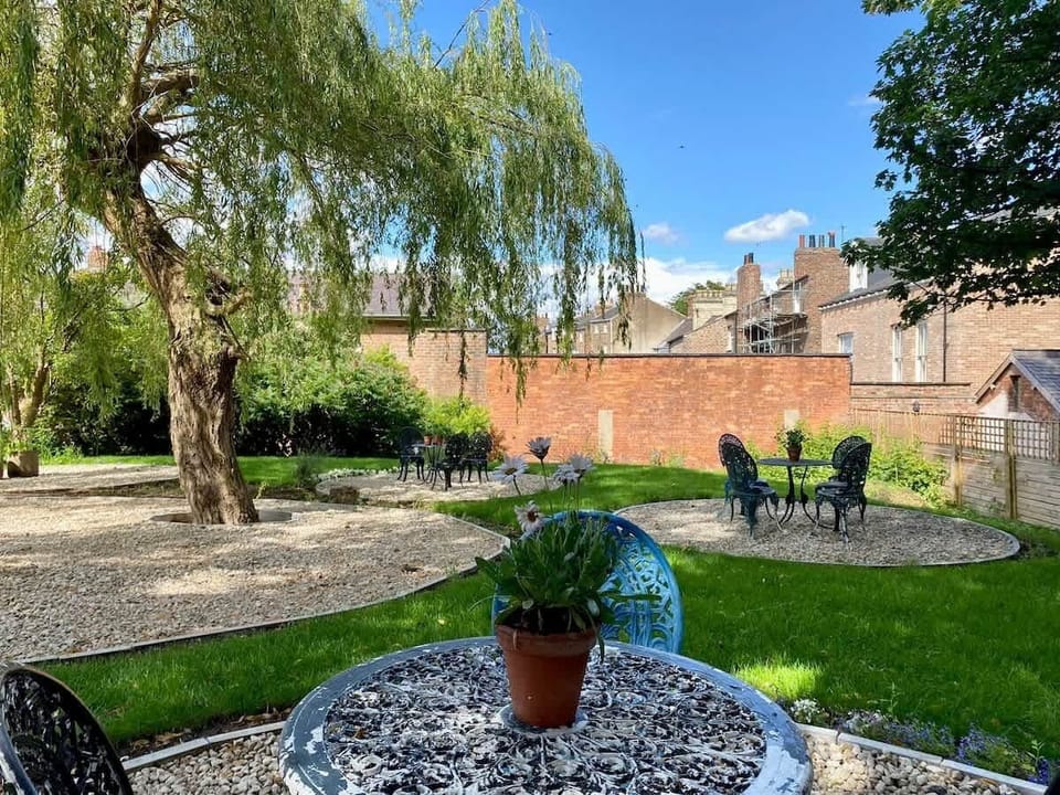 Tranquil garden with gravel paths and seating areas. A round metal table with a potted plant sits beneath a graceful willow tree, while matching chairs and decorative sections create a peaceful, inviting space framed by brick walls and greenery.
