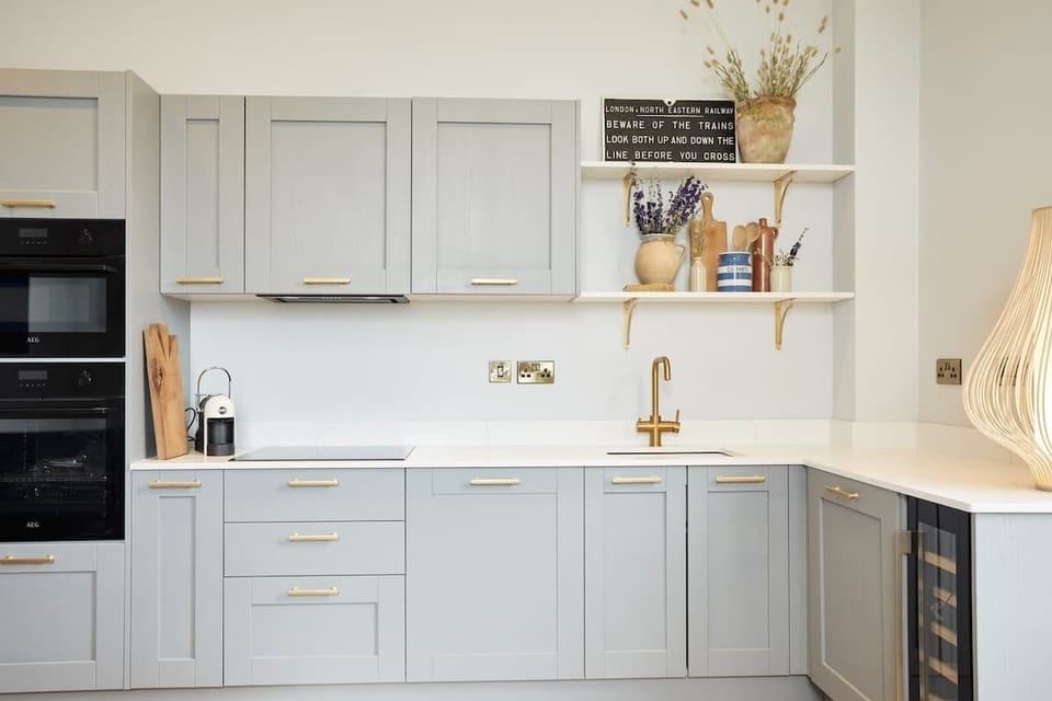 A sleek kitchen with gray cabinets, white counters, and built‑in oven. Gold faucet, floating shelves, and decorative touches create a warm, welcoming space filled with natural charm.