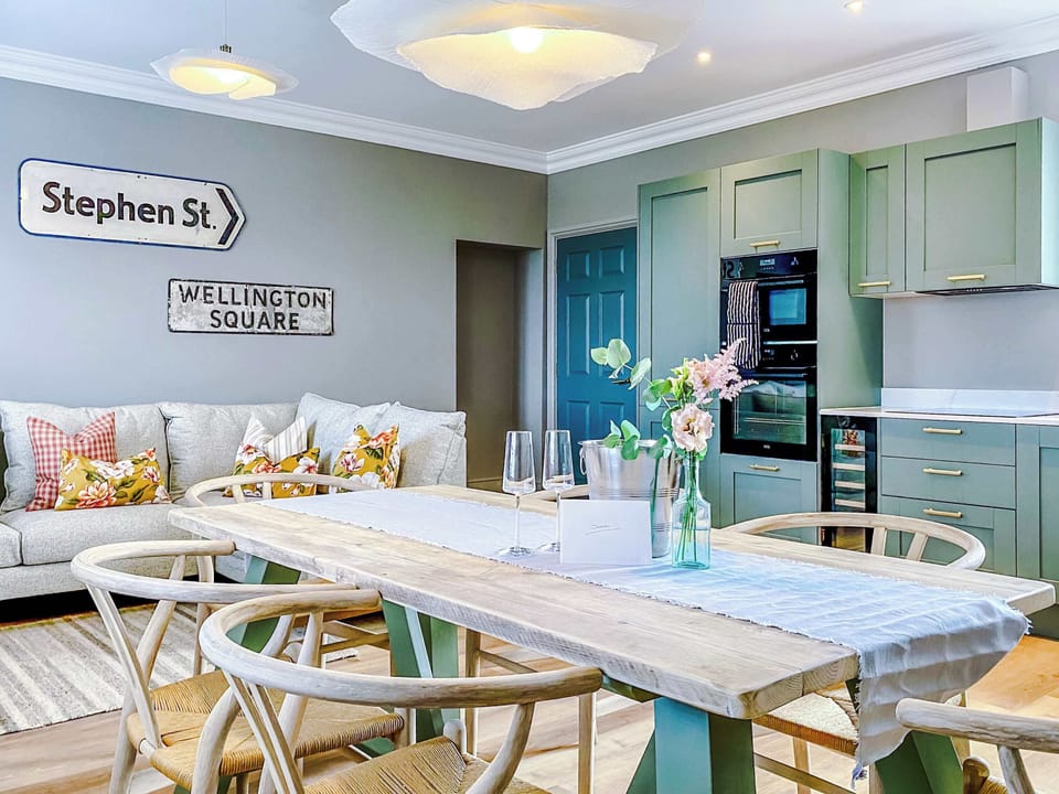Contemporary living room with a beige sofa layered in patterned pillows, set beneath decorative street signs for character. Warm lighting and cloud-like fixtures create a cozy, stylish space that flows seamlessly into the kitchen and dining area.
