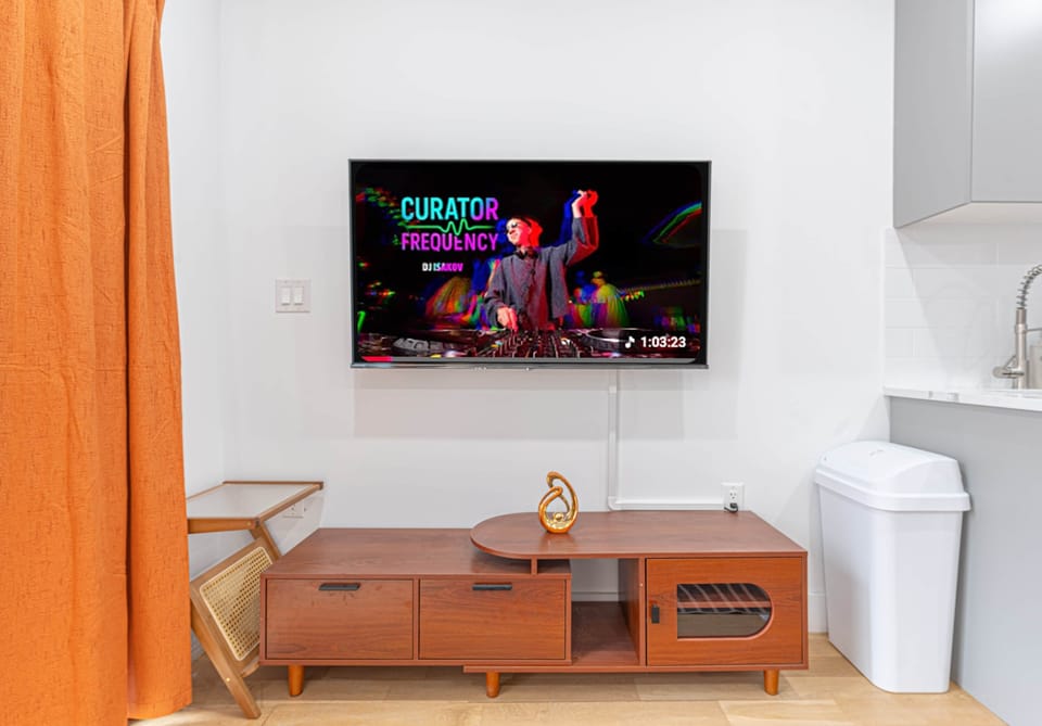 Wall-mounted TV above a wood console, next to orange curtains and a white bin.