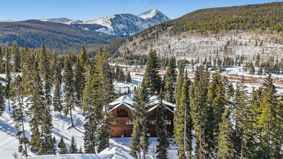 Mountain chalet nestled in snow-covered evergreen forest with dramatic alpine peaks creating a pristine winter wonderland setting.