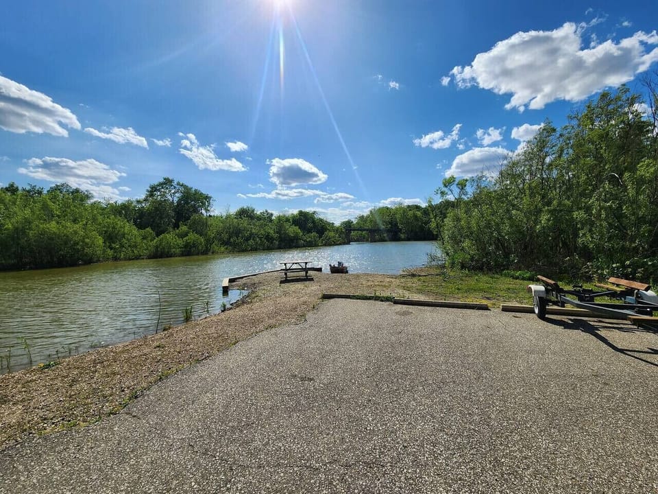 Private riverfront access with a boat launch, picnic table, and firepit under sunny blue skies. Surrounded by lush trees and calm water, this scenic waterfront spot is perfect for fishing, kayaking, relaxing, and enjoying nature.
