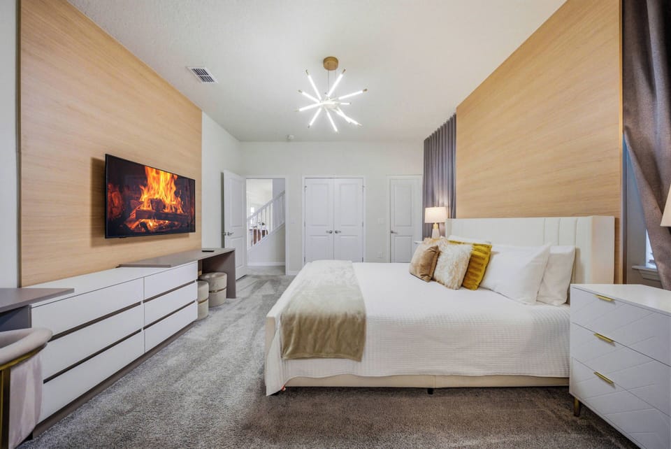 Welcome to a stylish bedroom with a plush king-sized bed dressed in soft linens and inviting pillows. Warm wood paneling and sleek white furniture create harmony, while a modern chandelier adds a sophisticated touch for your stay.