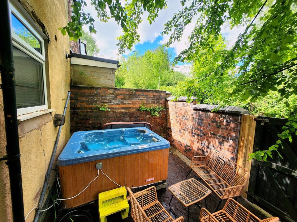Outdoor patio area with a hot tub, brick walls, and metal seating beside a gate and surrounding trees.
