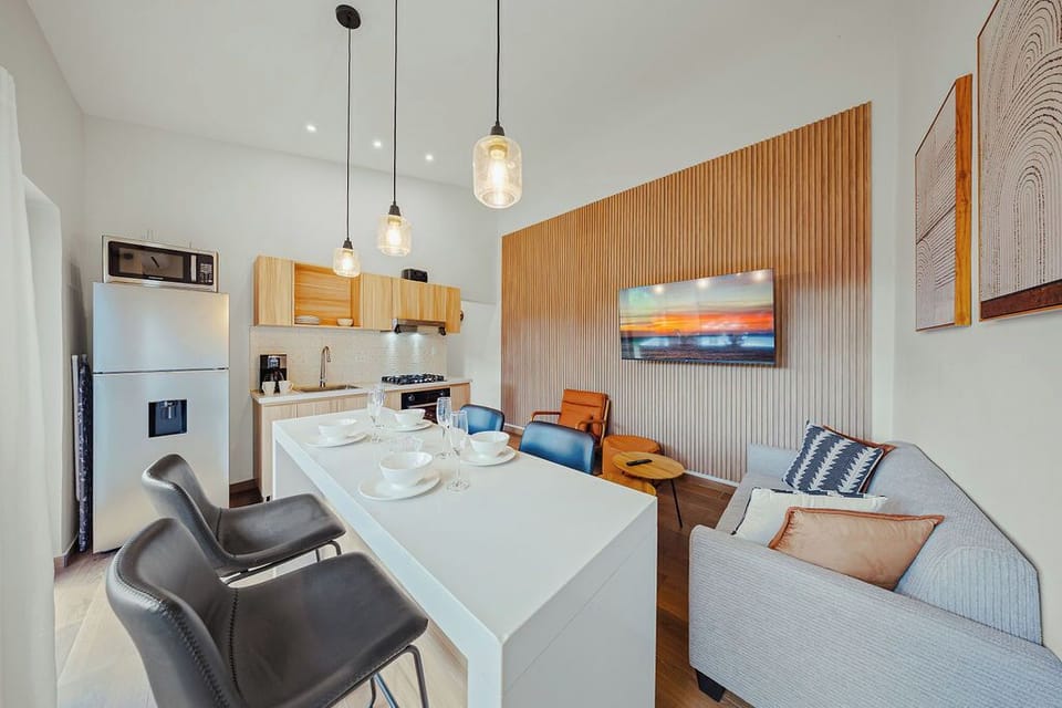 Stylish open-plan kitchen and lounge with wood accents, pendant lighting, and cozy seating. Features a sleek island, stainless steel appliances, and a slatted TV wall. Artful touches and warm tones create a welcoming, modern vibe.