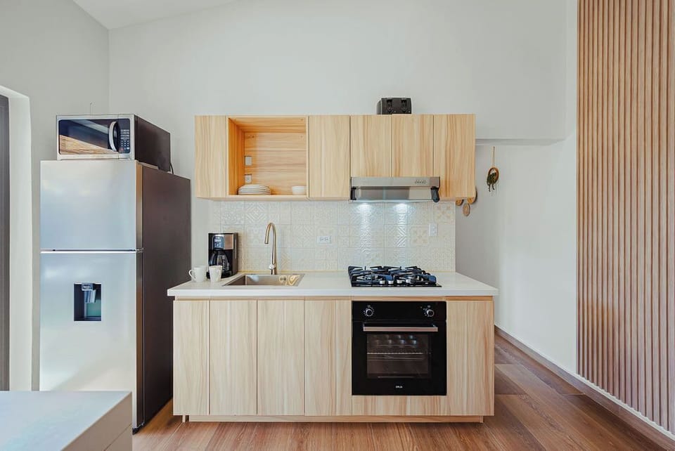 Minimalist kitchen with light wood cabinetry, white countertops, and stainless steel appliances. Features a gas cooktop, built-in oven, and sleek range hood. Warm wood flooring and slatted accent wall add texture to the clean, modern design