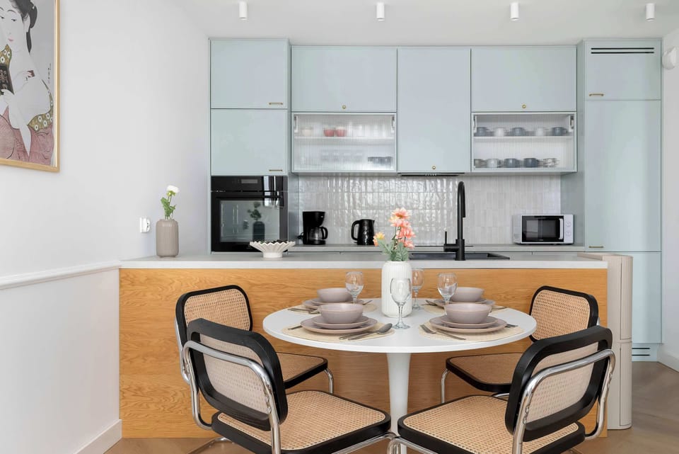 A modern kitchenette and dining area showcase light blue cabinetry and a round wooden table. Elegant black-framed chairs and a designer pendant lamp create a perfect spot for meals.