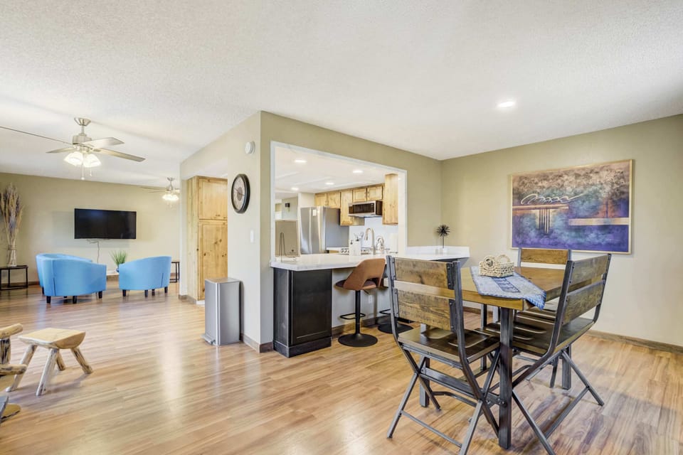 The dining space features a high-top wooden table with seating for four, positioned between the kitchen and living area. Large windows and a modern ceiling fan keep the space bright and airy for group meals in the home.