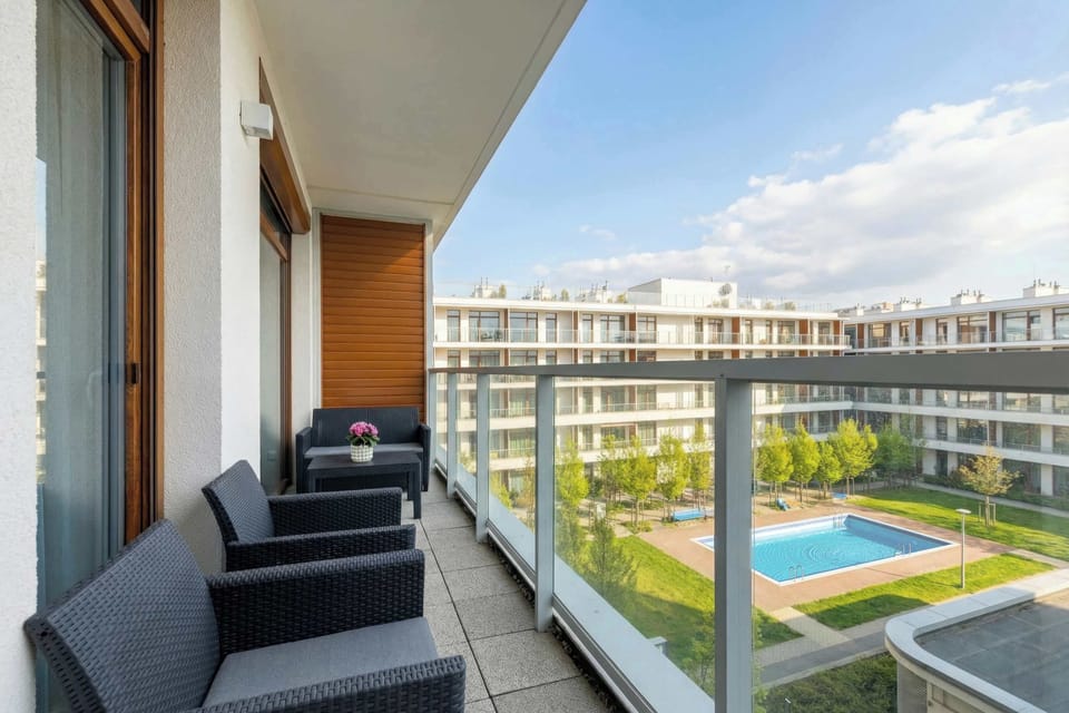 A balcony furnished with outdoor seating overlooking the pool and courtyard. Glass railings provide an unobstructed view of the complex. The space is suitable for relaxing outdoors.