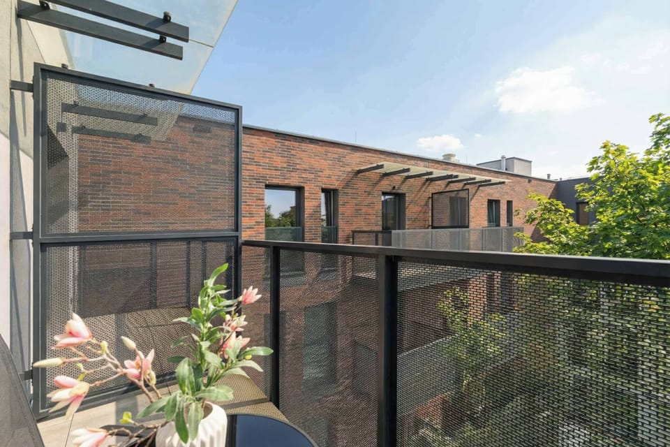 The balcony view shows the glass railing and a small table decorated with pink flowers. Beyond the terrace, the architecture of neighboring brick and plaster buildings is visible.