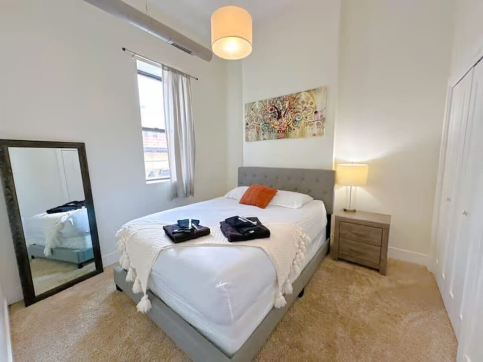 A cozy bedroom with a soft upholstered bed, warm lighting, and a large mirror leaning against the wall.
