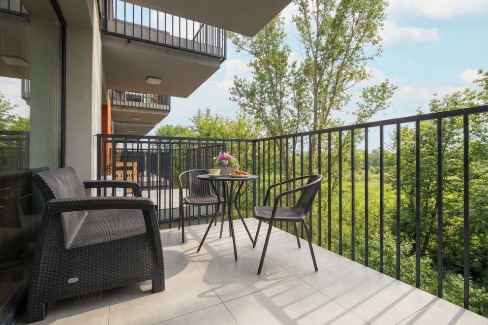 A balcony furnished with outdoor seating and a small table. Black railings frame the space, overlooking green trees. The area appears suitable for relaxing outdoors.