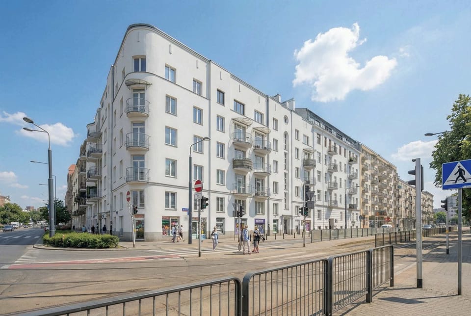 An exterior view of a modern residential building with balconies and large windows under a blue sky. A street and tram tracks run in front of the property.