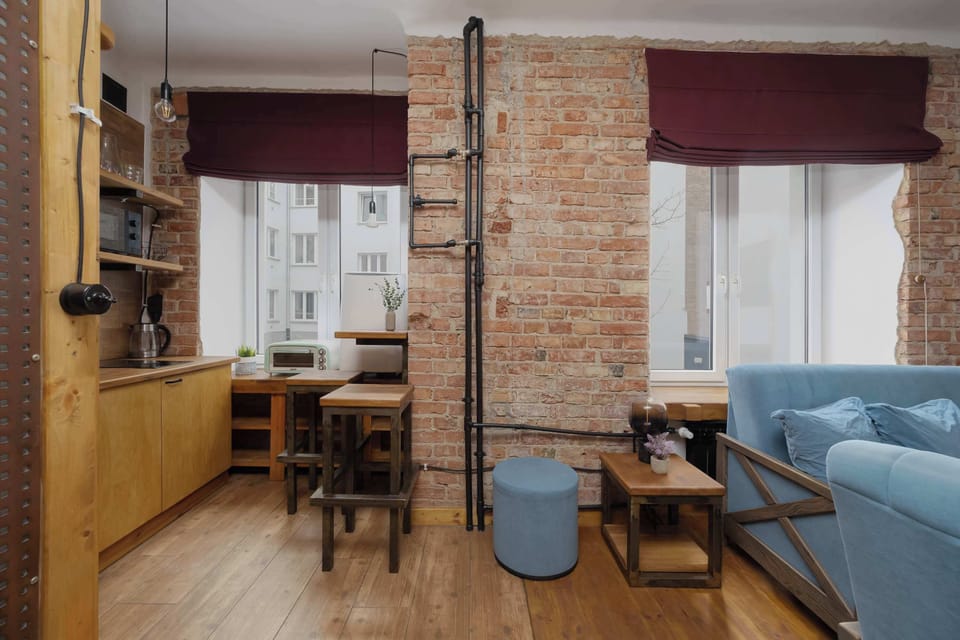 A wider view of the open-plan living room showing a small dining table, blue sofa, and brick pillars dividing the space. Burgundy curtains and wooden flooring tie the room together.