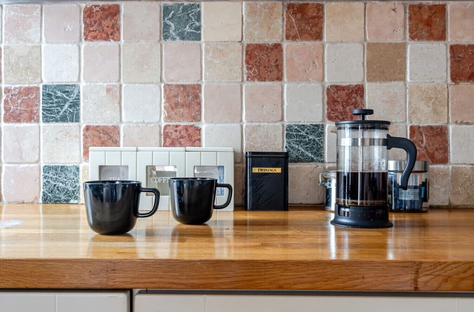 Kitchen worktop set out with mugs, a French press, a tea canister and tiled splashback behind.