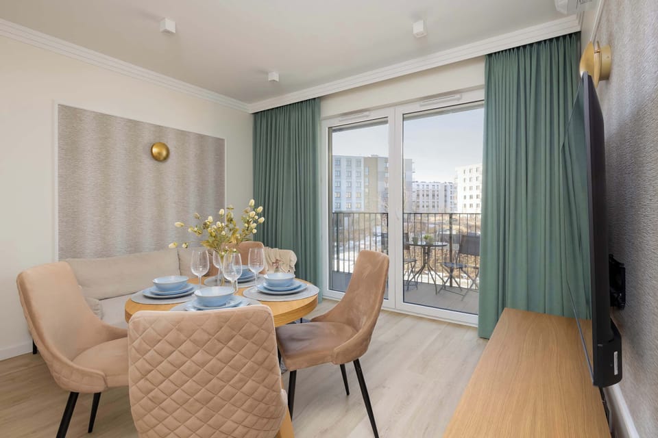 A round table set for four stands in a sunlit corner near balcony doors, decorated with a vase and elegant tableware.