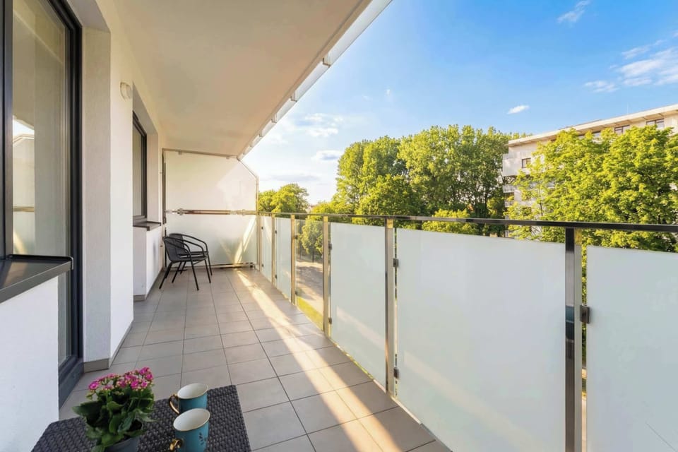 The private balcony is equipped with a small table and chair, offering an outdoor space to enjoy the fresh air. Glass railings provide an unobstructed view of the surrounding greenery and street.