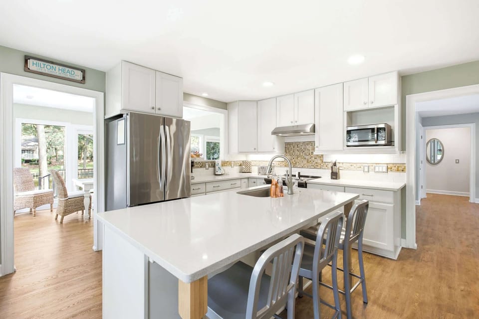 Crisp white cabinetry and cool stone countertops create a bright, airy hub for morning coffee and shared meals. Soft light pours across the warm wood floors, highlighting a modern kitchen designed for effortless hosting and culinary ease.
