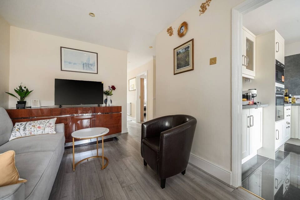 A space to gather in the living room, with a grey sofa, wall-mounted TV on a wooden sideboard, and an open doorway leading through to the kitchen.