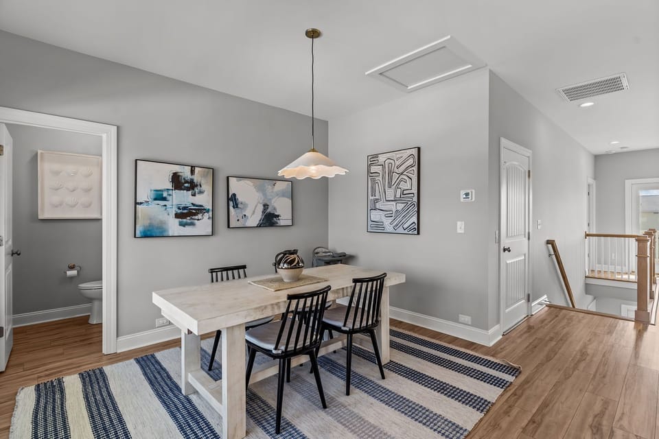 Bright Oak Island NC dining area with wood table seating, coastal artwork, large windows, and glass door leading to the covered balcony and outdoor seating.