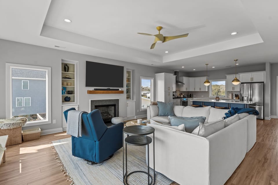 Spacious open-concept living room and kitchen with sectional sofa, fireplace, large TV, coastal décor, and bright kitchen with island seating in this Oak Island, NC beach home.
