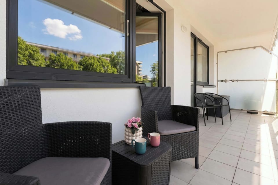 A spacious balcony furnished with dark wicker chairs and a small table. The tiled floor and glass railing create a modern outdoor setting. Green trees are visible beyond the balcony.