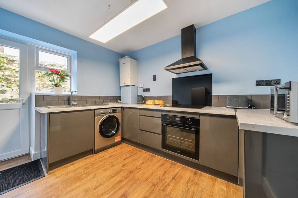 Grey fitted kitchen with a built-in oven, washing machine, extractor hood, white worktops, and subway-tile splashback, with a window to one side and blue-painted walls.
