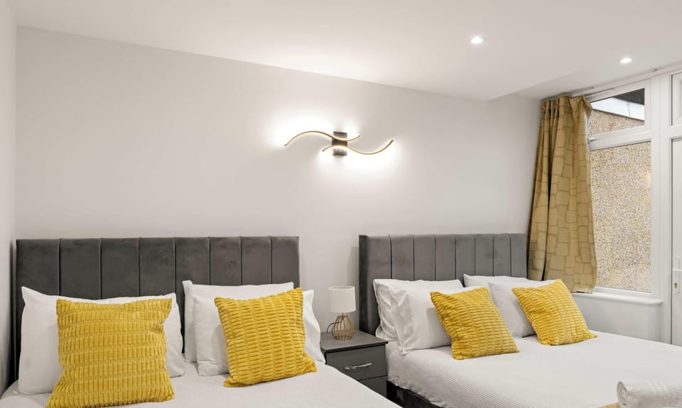 Twin beds with dark grey headboards and vibrant mustard-yellow accents sit symmetrically around a slim bedside table. A sleek, curved wall sconce provides warm lighting against the room’s crisp, white walls.