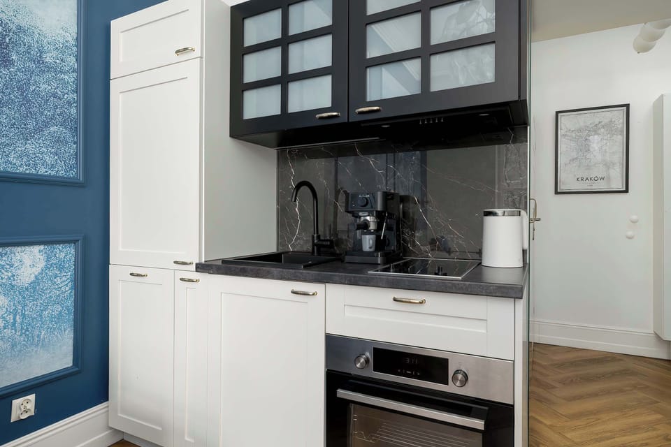 A compact kitchenette with white cabinetry, wooden flooring, and black glass-front upper cabinets. A built-in oven and stovetop are neatly integrated. The space looks clean, functional, and modern.