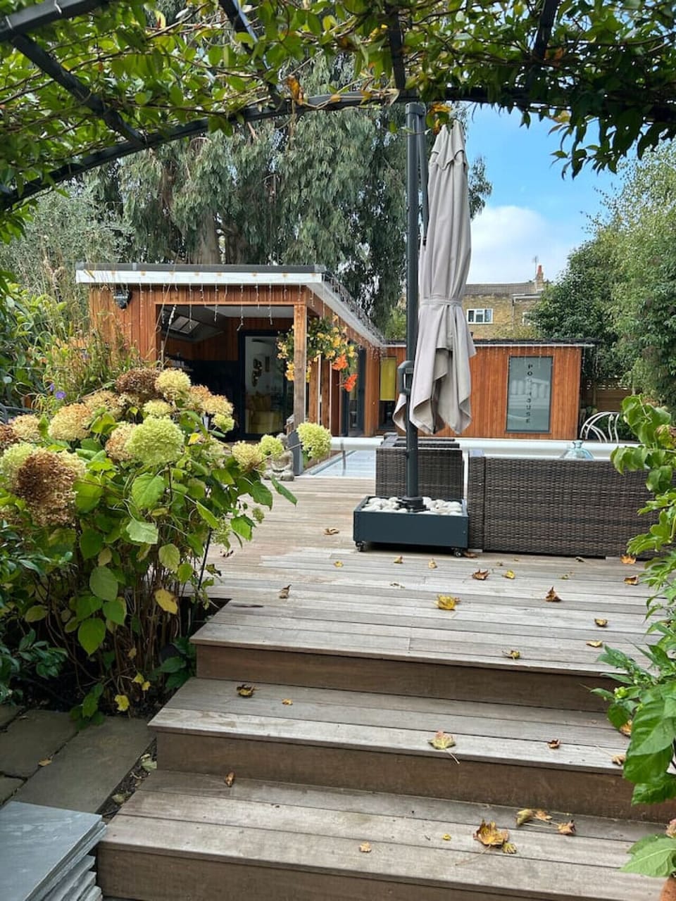 A stepped wooden decking path winds through the garden from the gate area towards the pool house entrance, bordered by mature plants and flowering shrubs.