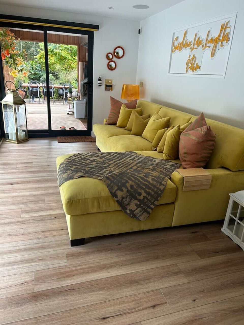The living area with the large yellow corner sofa, room-darkening roller blind lowered, and the glazed patio doors open to the garden beyond.