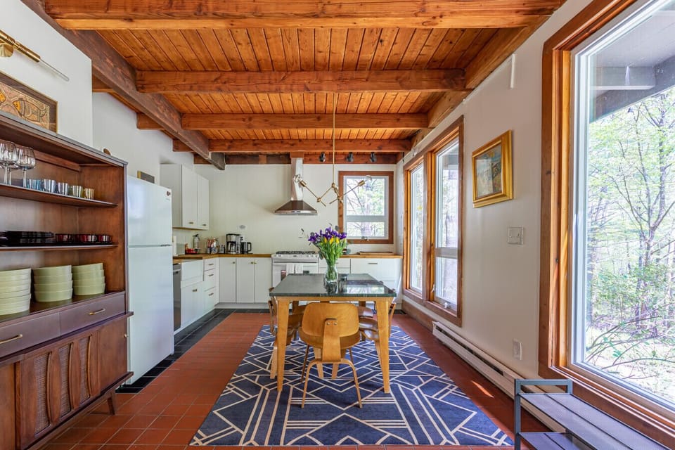 This sunlit kitchen and dining area is surrounded by oversized windows overlooking peaceful woodland scenery
