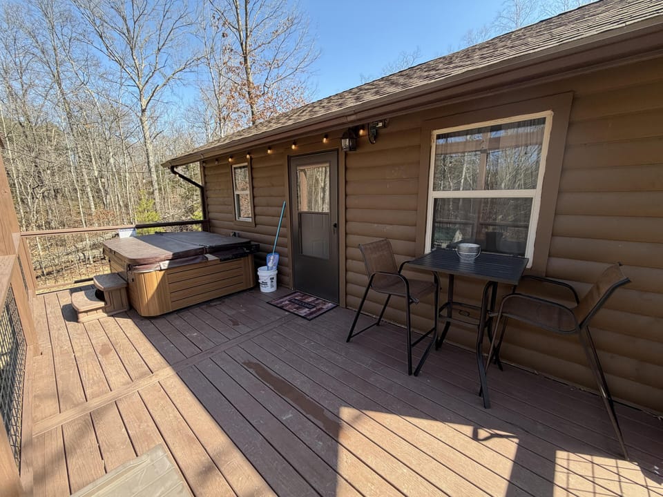 Wood-plank deck features a covered hot tub, two brown mesh chairs, and a black metal table with a bucket.