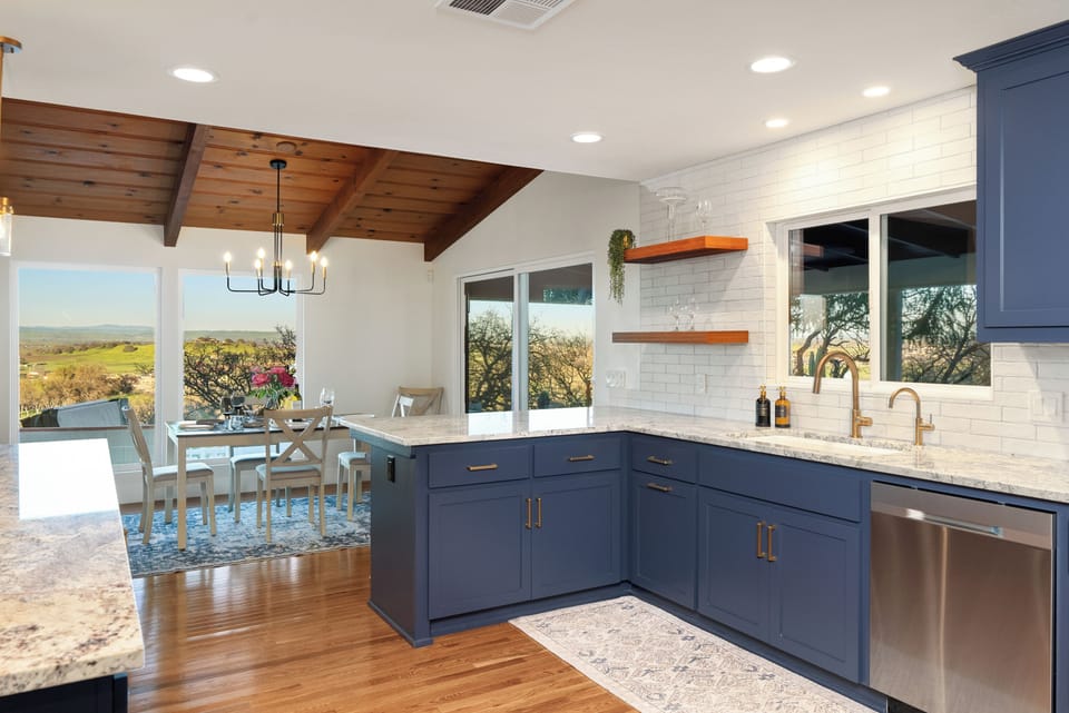 A modern navy kitchen featuring granite countertops and open wood shelving flows into a bright dining area with vaulted wood-beamed ceilings and large picture windows that frame sweeping views of the rolling green hills.