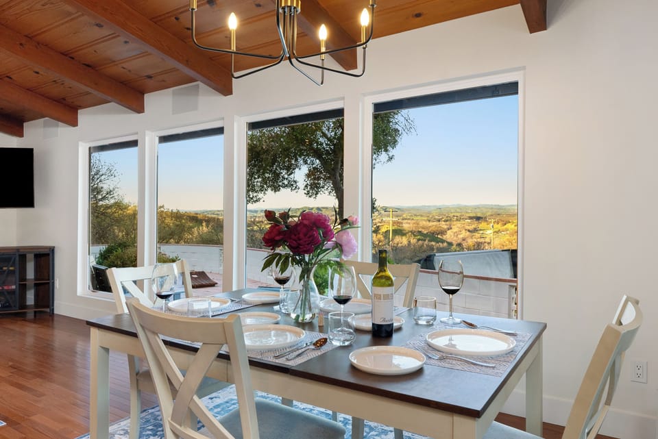 A warm, open-concept dining area featuring a set table with fresh flowers, vaulted wood-beamed ceilings, and large picture windows that frame sweeping views of the rolling hills, perfect for sunset dinners and entertaining.