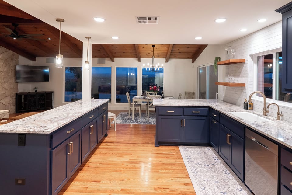 A modern navy kitchen features elegant granite countertops, wooden open shelving, and warm hardwood floors that flow into an open-concept dining area with vaulted wood ceilings, perfect for a seamless indoor-outdoor living experience.
