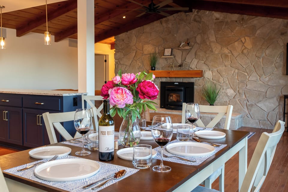 A modern navy kitchen with stone counters and a warm dining area that flows into a living space highlighted by a floor-to-ceiling stone fireplace and vaulted wood-beamed ceilings.
