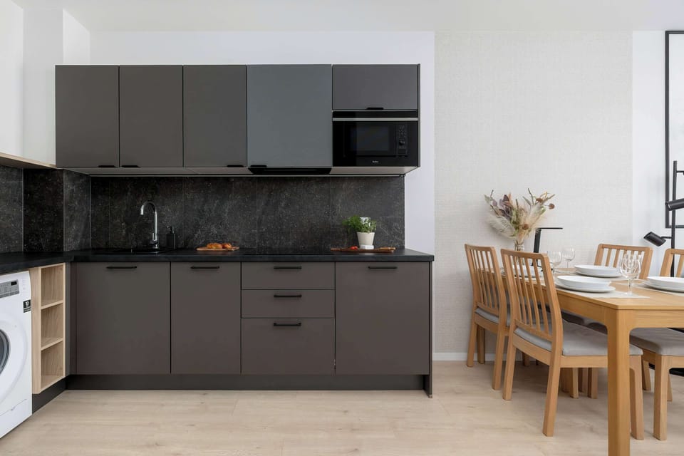 A sleek kitchenette features dark grey cabinetry, integrated appliances, and a wooden dining table that seats four. The contrast between the dark furniture and light walls adds a touch of contemporary elegance.