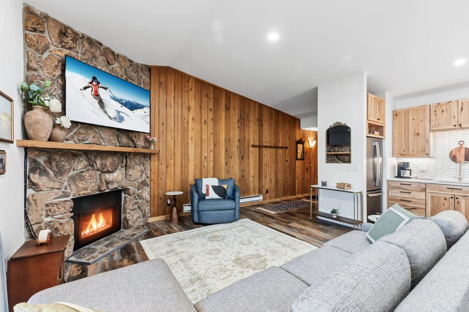 Cozy up by your crackling stone fireplace while watching the big screen, with warm wood details creating the perfect mountain retreat atmosphere.