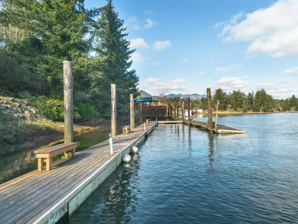 Private dock extends into pristine waters, surrounded by towering evergreens and distant mountain peaks under cloudy skies.