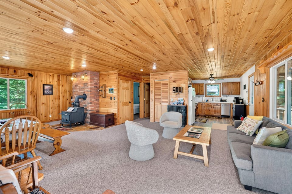 Large open living area with a gray couch, wood coffee table and two gray swivel chairs. In the background is a wood stove.