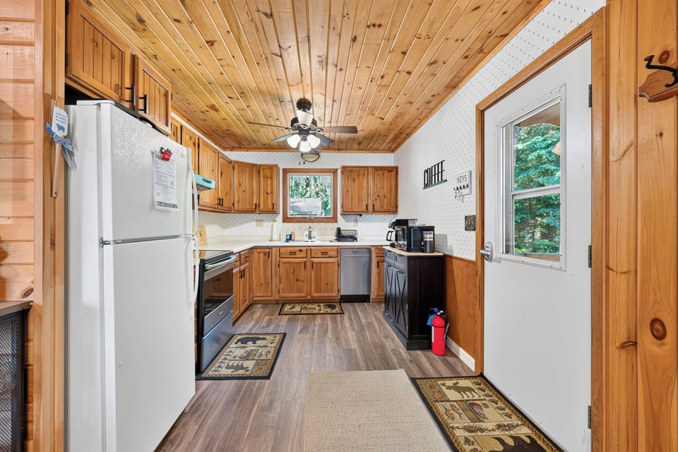 There is a white refrigerator, a black stove and a stainless steel dishwasher, with wood cupboards and a wood floor. There is a black cupboard for coffee accessories.