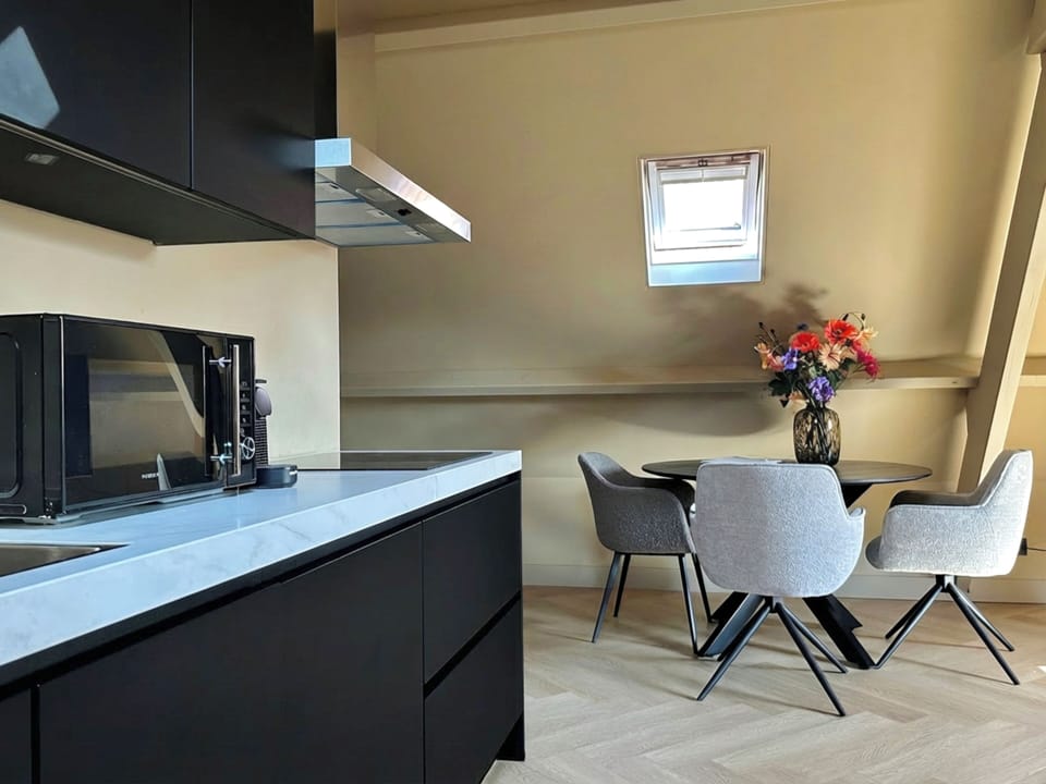 Modern black kitchen with sleek countertop and coffee machine, opening to a cozy dining area with round table, chairs, skylight window and decorative flowers.
